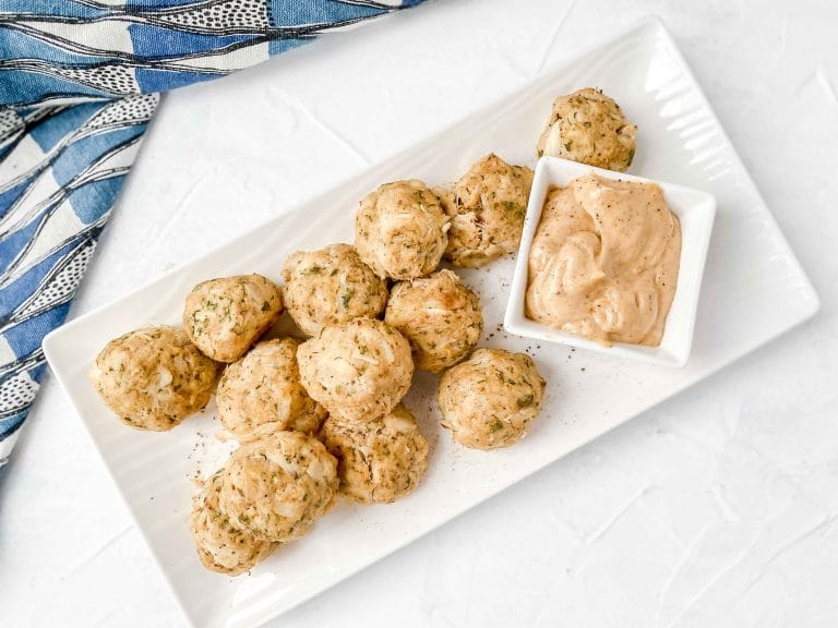 Image is a top angle view of Paleo Maryland Crab Ball on a white rectangle plate. There is a bow of Old Bay Aioli on the plate. The plate is on a gray background with a black and white dish towel on the left. www.atwistedplate.com/paleo-maryland-crab-balls/