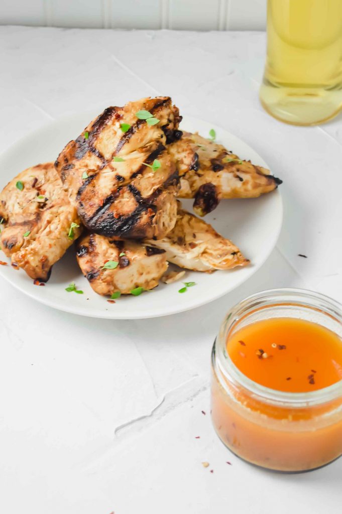 Image for BBQ and Mustard Chicken Marinade.  In the bottom right there is a jar of the Marinade.  In the middle left of the image are four pieces of grilled chicken on a white plate topped with parsley.  In the top left there is the a jar of olive oil.  https://www.atwistedplate.com/bbq-and-mustard-chicken-marinade/