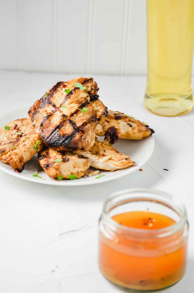 Image for BBQ and Mustard Chicken Marinade.  In the bottom right there is a jar of the Marinade.  In the middle left of the image are four pieces of grilled chicken on a white plate topped with parsley.  In the top left there is the a jar of olive oil.  https://www.atwistedplate.com/bbq-and-mustard-chicken-marinade/