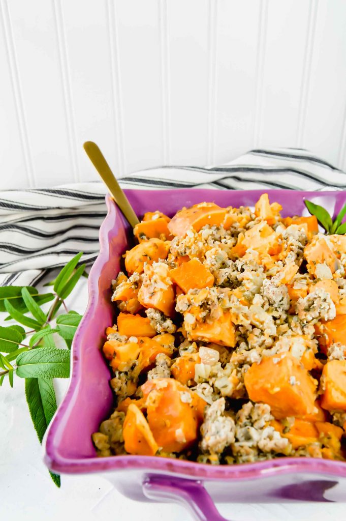 Image: angled view of Sweet Potato Stuffing in a purple dish.  Around the dish are fresh sage leaves.  A black and white towel is at the top of the image.   https://www.atwistedplate.com/sweet-potato-stuffing/