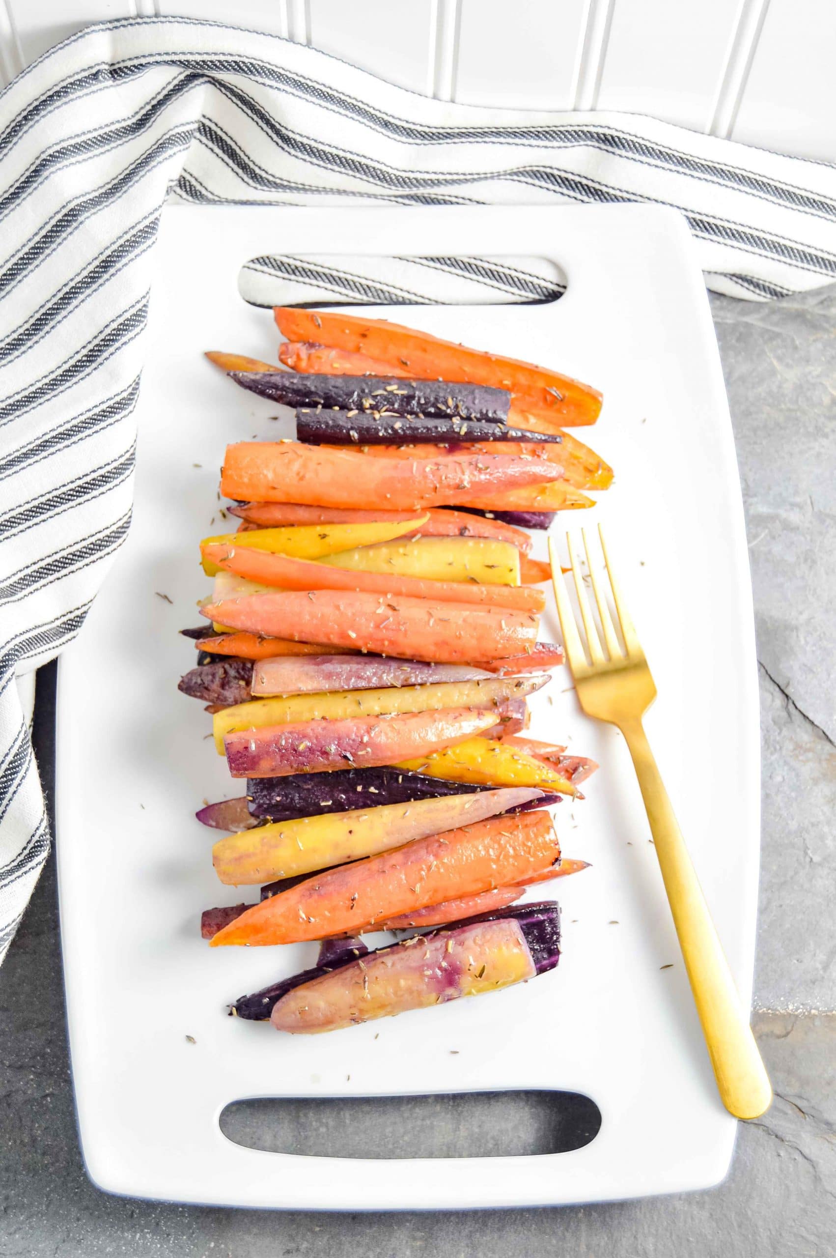 Image of a side angle view of Perfectly Roasted Carrots on a rectangle white plate with a gold fork.  The plate sites on a gray blue background with a black and white striped towel in the top left corner.   https://www.atwistedplate.com/perfectly-roasted-carrots/