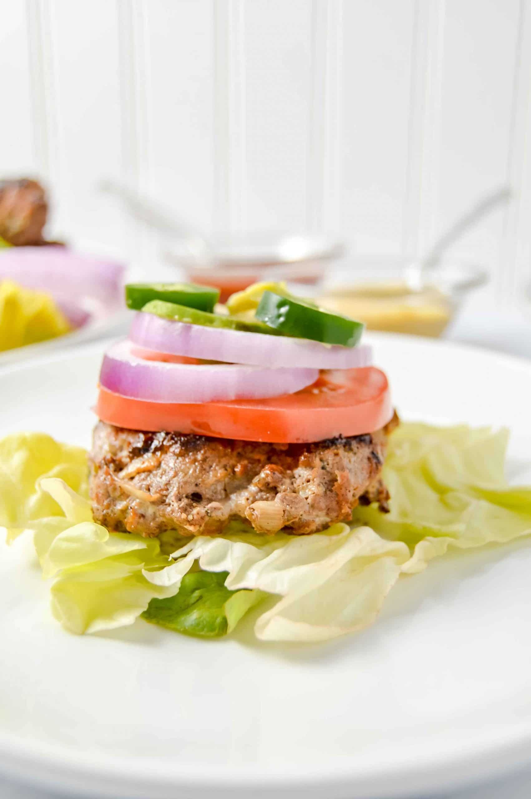 Image is a side view of a Ranch burger on top a bed of lettuce with a round white plate. Atop the burger red onion, tomato, mustard, ketchup, jalapeño and avocado. There is a bowl of mustard and a bowl of ketchup above the plate. Next to it is a cut off plate with burger and red onion. www.atwistedplate.com/savory-ranch-burger/