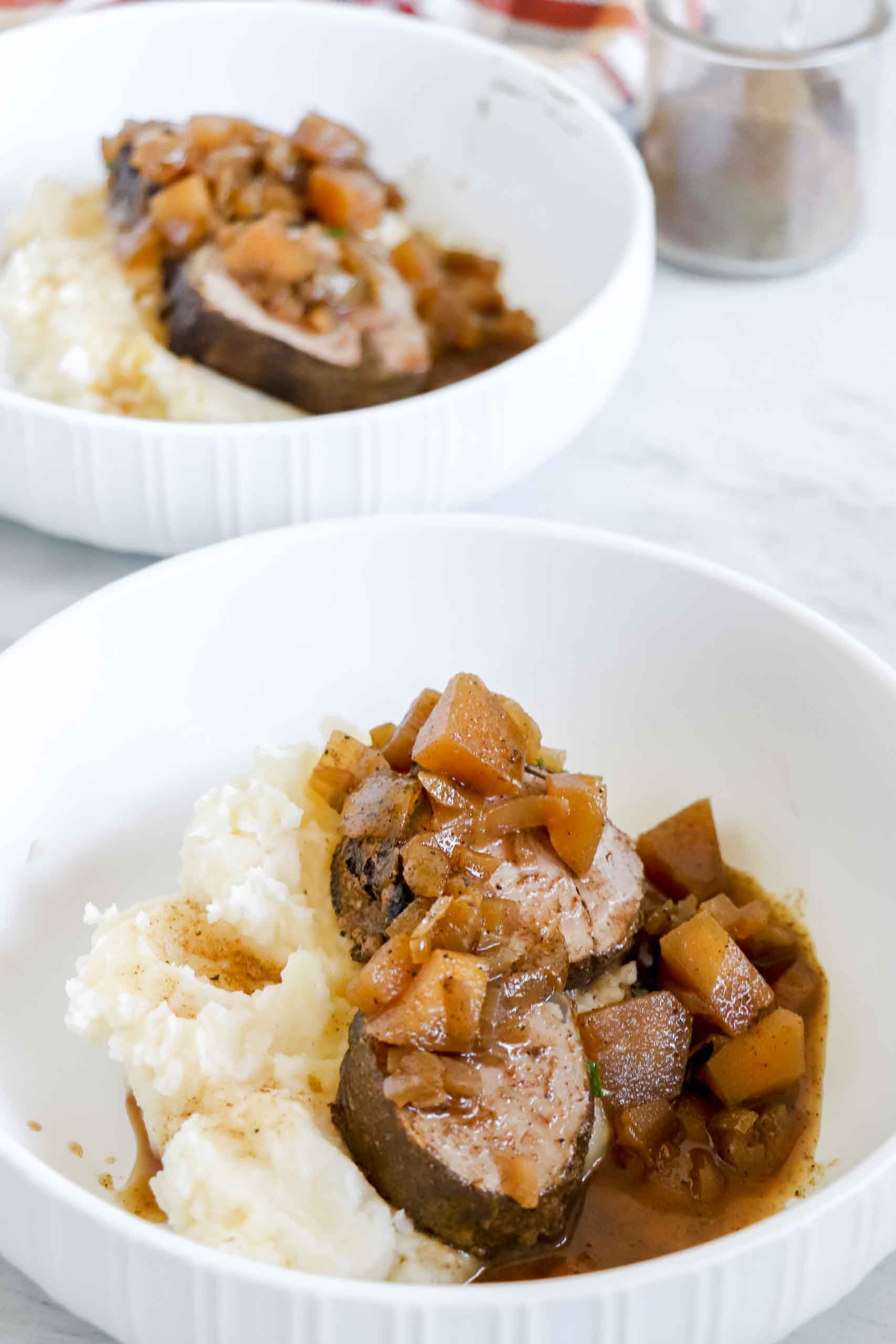 Pork tenderloin with Mashed Potatoes on a white plate with a plate of pork tenderloin with apples in the background. https://www.atwistedplate.com/pork-tenderloin-with-apples
